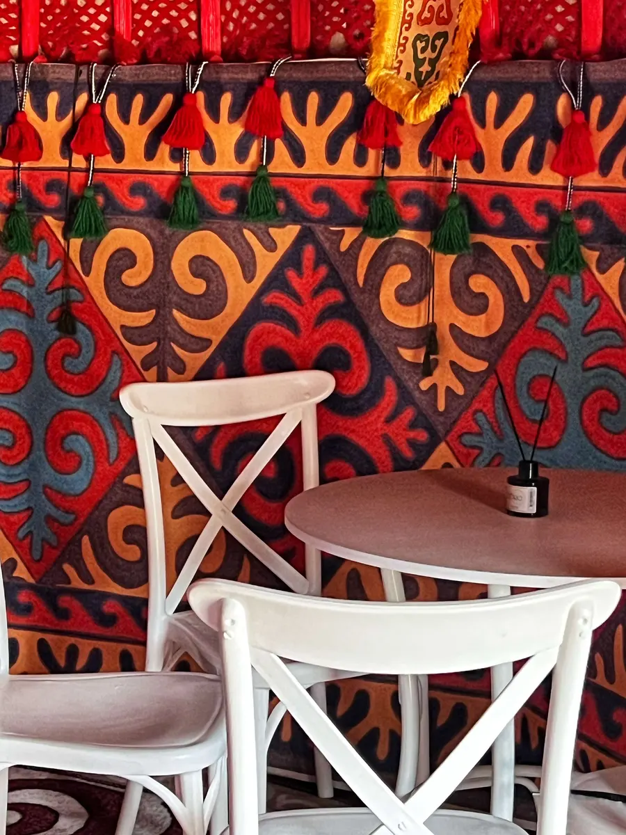 White wooden chairs and a table set against a yurt wall with rich Kazakh textile patterns